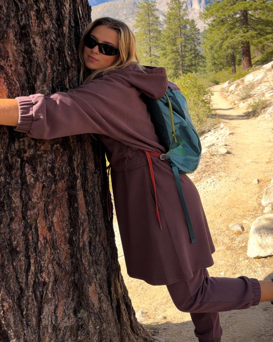 Person in a purple outfit with a blue backpack hugging a tree in a natural setting