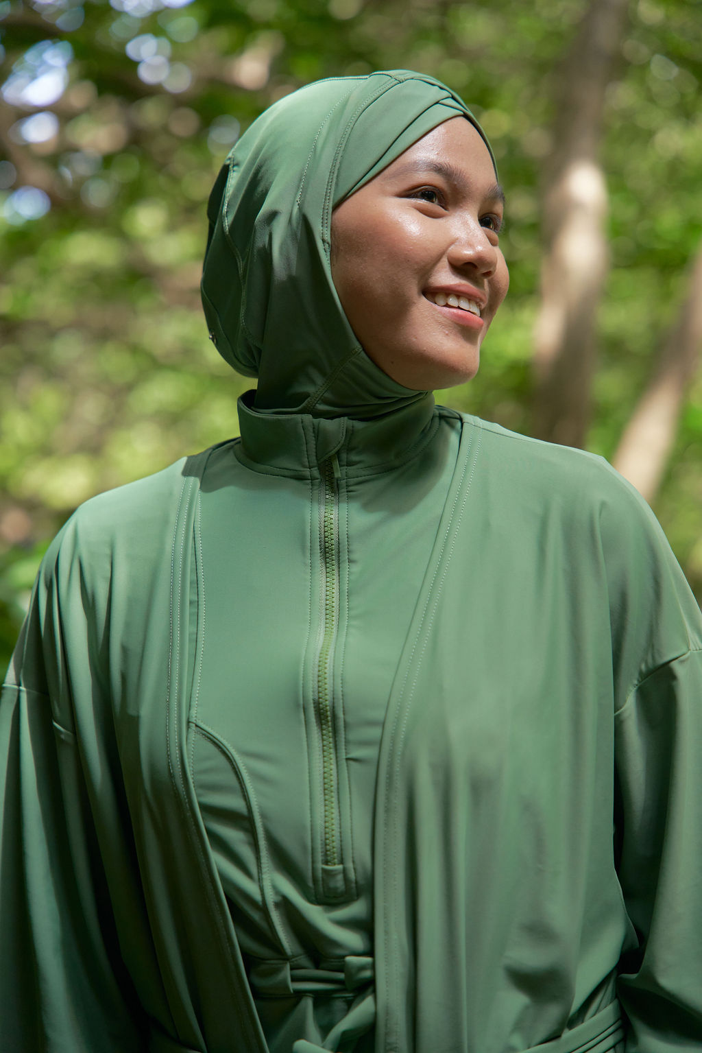 A woman wearing a matching olive green hijab and lightweight zip-front abaya or long top, photographed outdoors with a blurred forest background.
