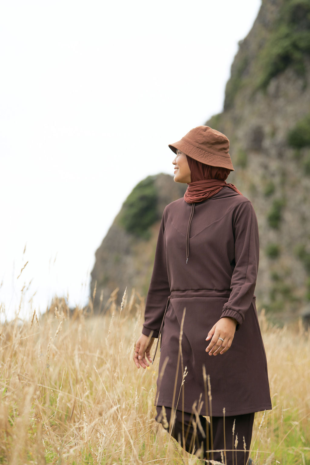 Person wearing a brown outfit and hat standing in a field with mountains in the background