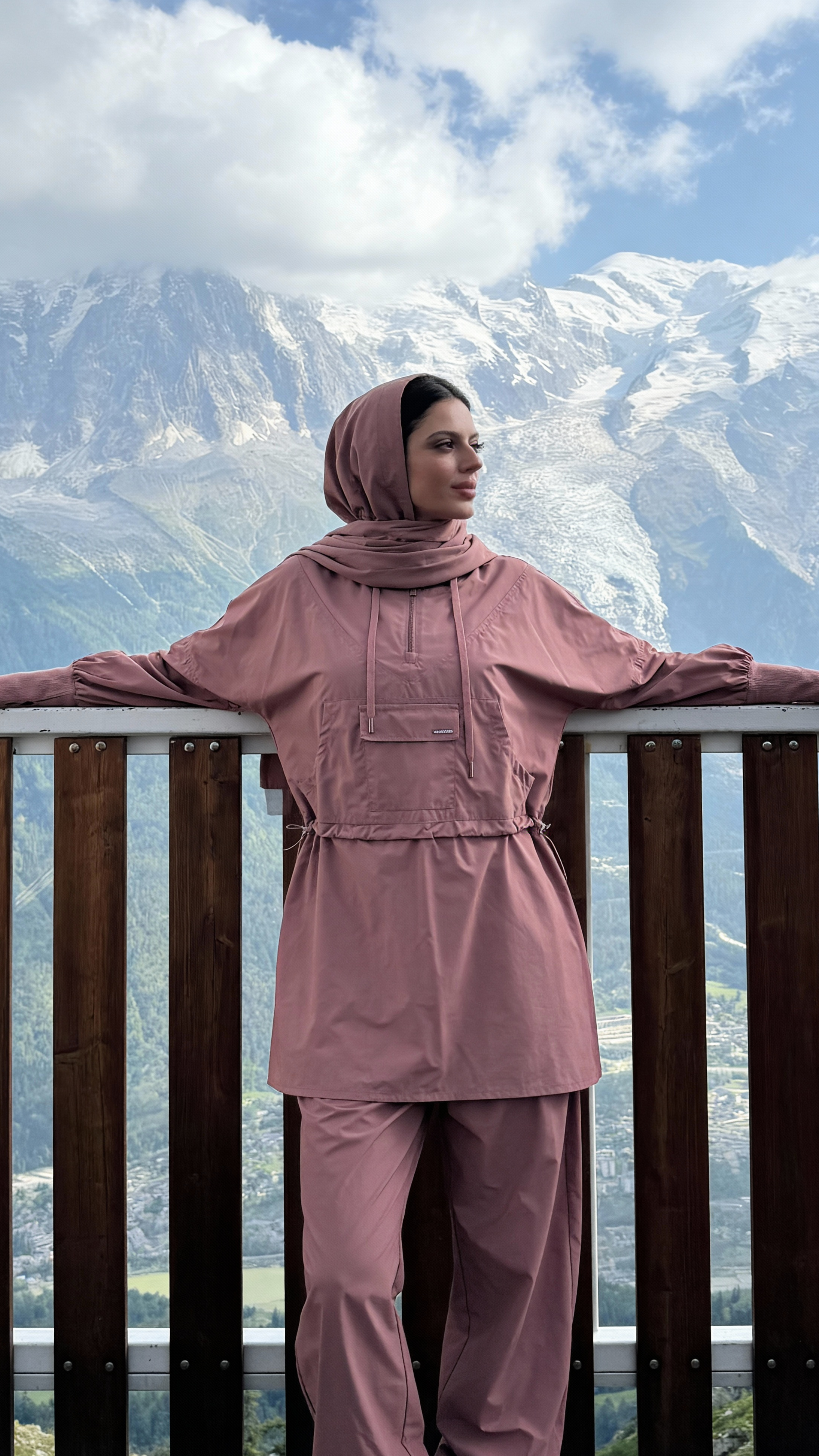 Person wearing a purple outfit with a hijab standing against a mountainous landscape.