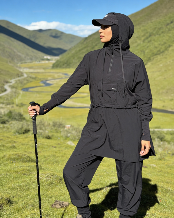 Person wearing a black outdoor outfit with a hood, standing in a scenic mountain landscape.