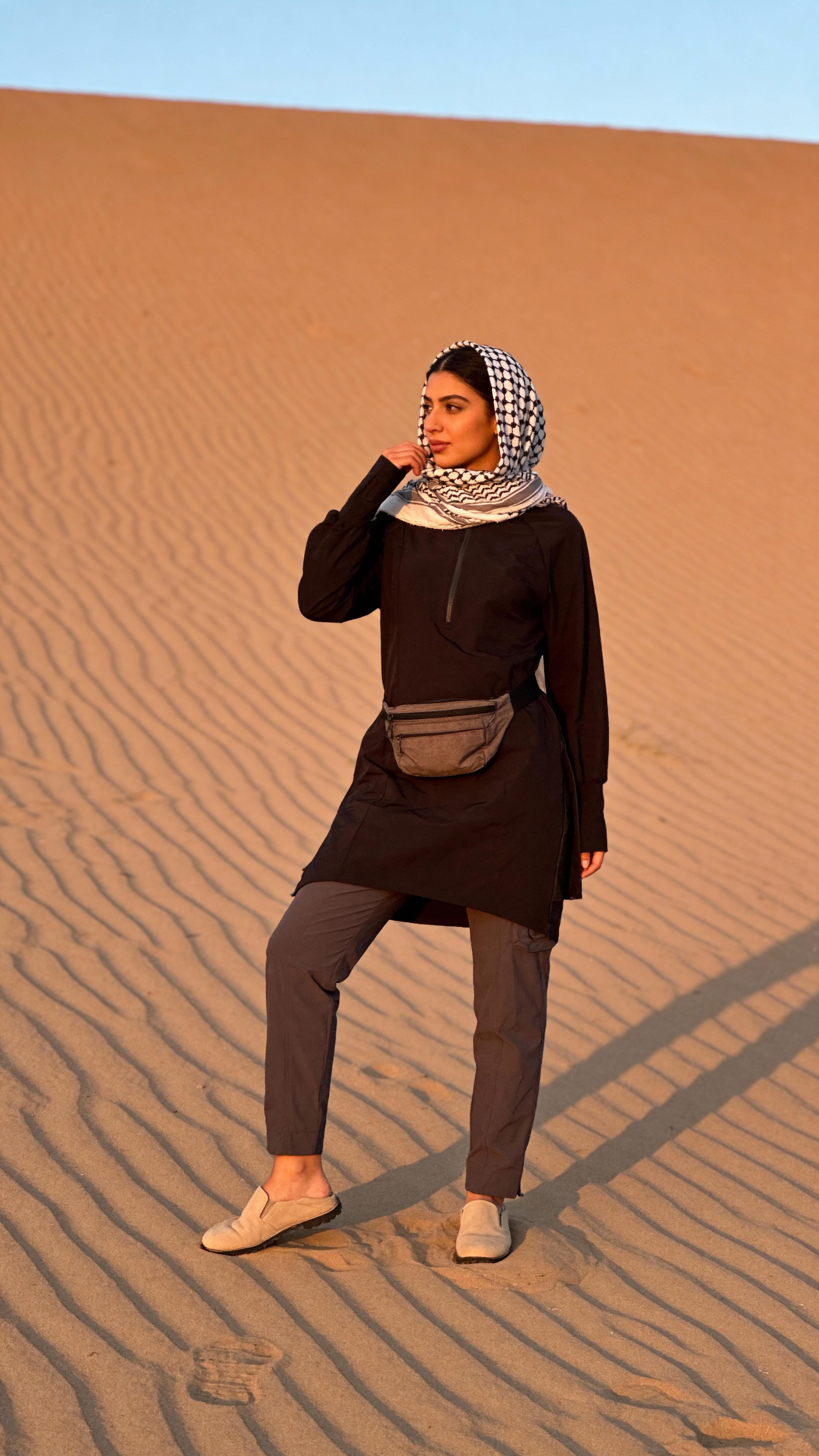 Person standing in a desert landscape wearing a black outfit and headscarf.