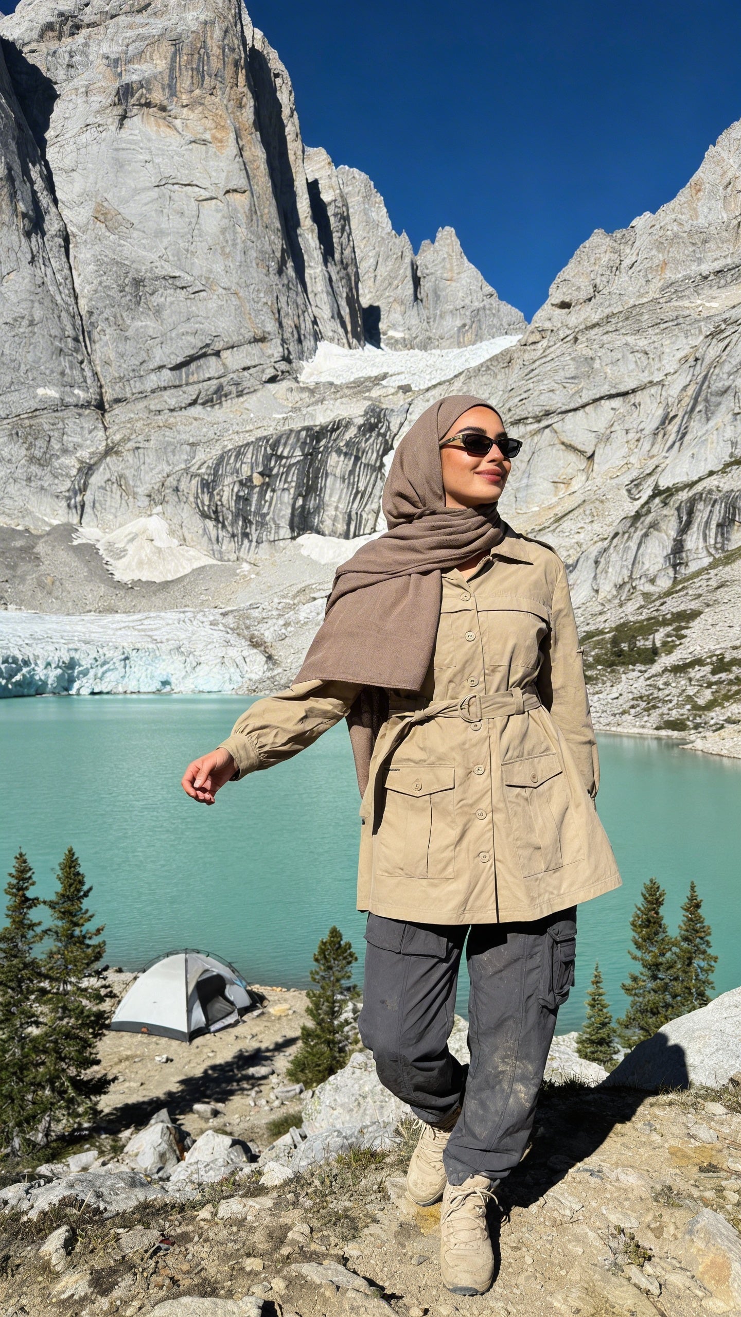 Person in beige jacket and hijab standing in a mountainous landscape with a lake and tent.