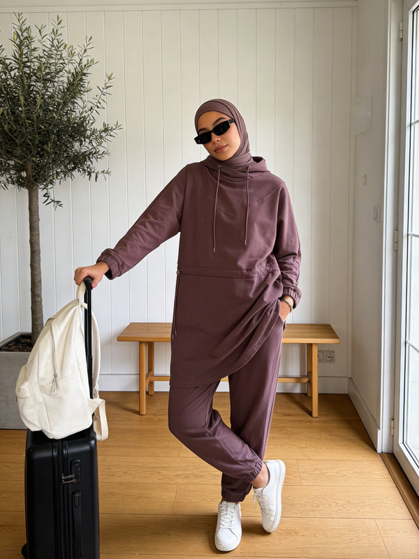 A woman wearing a matching dusty mauve hijab and two-piece lounge set consisting of a long-sleeve hooded sweatshirt and jogger pants, posing with a black suitcase and a white backpack in a bright room with wooden floors and a bench.