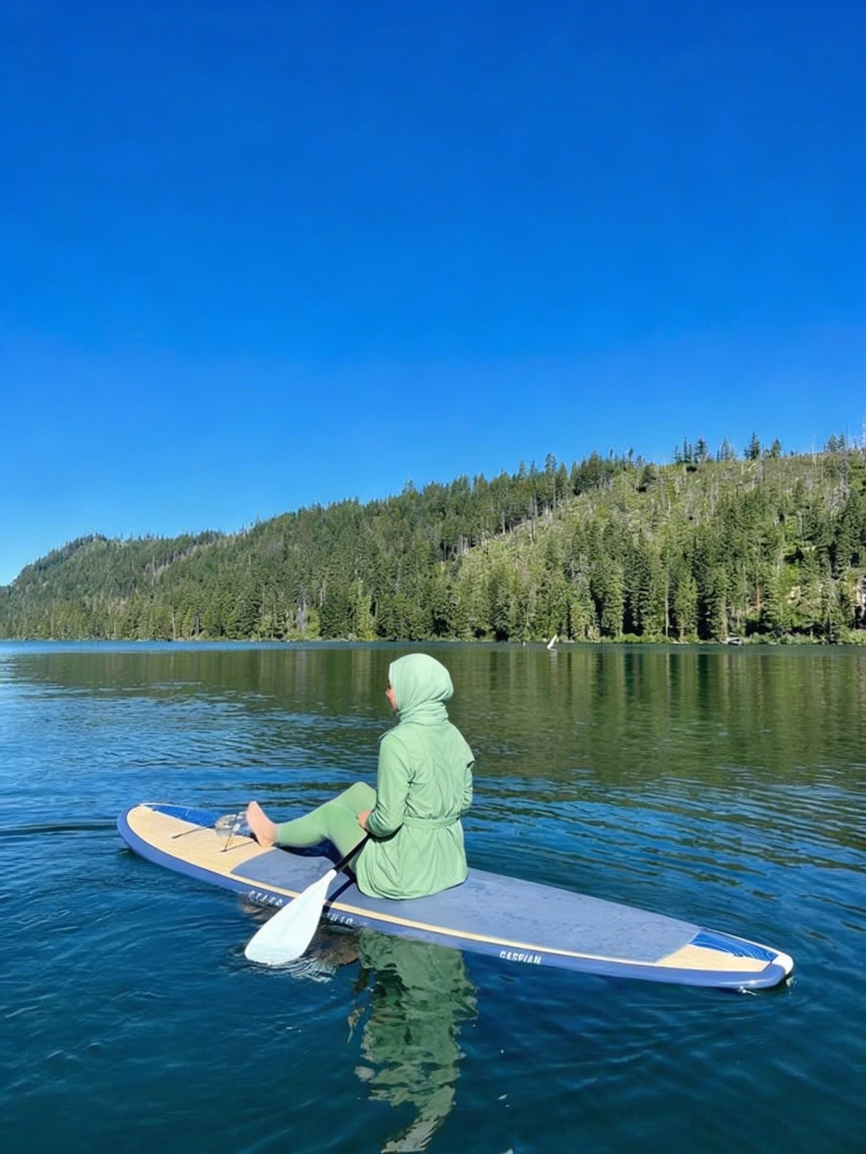 Person paddleboarding on a lake with a clear blue sky and forested shoreline.