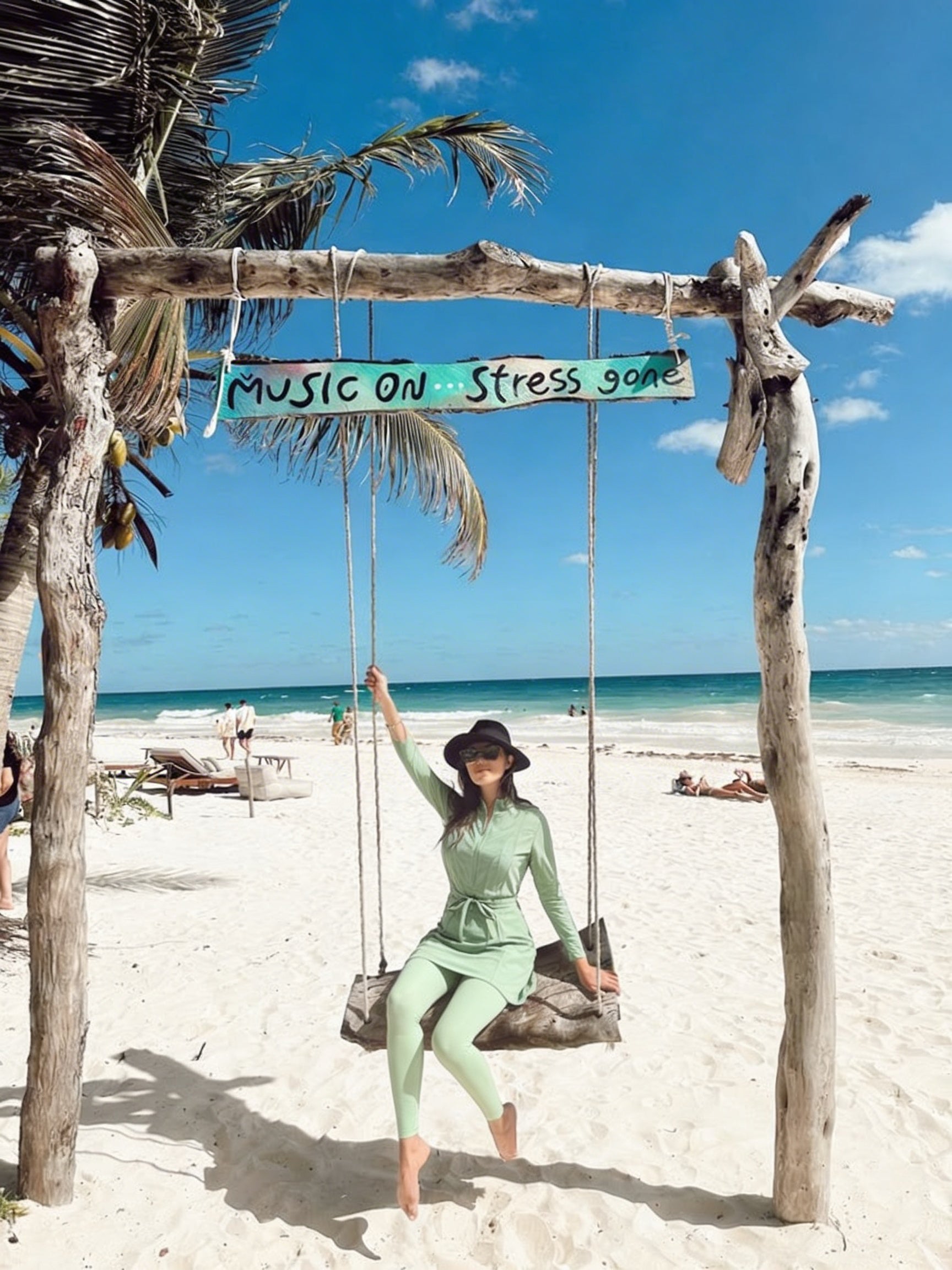 Woman on a swing with 'Music on Stress gone' sign at a tropical beach.