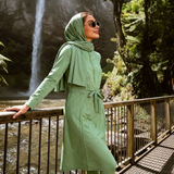A woman wearing a sage green matching hijab, long-sleeve tunic, and leggings stands on a metal walkway beside a waterfall in a forest setting.