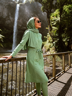 A woman wearing a sage green matching hijab, long-sleeve tunic, and leggings stands on a metal walkway beside a waterfall in a forest setting.