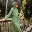 A woman wearing a sage green matching hijab, long-sleeve tunic, and leggings stands on a metal walkway beside a waterfall in a forest setting.